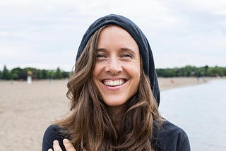 Photo of the interview subject, Jo-Anne McArthur, smiling widely on a beach, wearing a navy hoodie