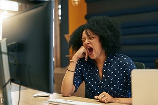 A young woman yawning at her desk while looking at her computer.