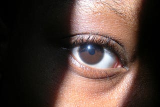 Closeup of a Black woman’s eye, with light shining in a strip where her eyes is. Rest of face obscured by shadow.