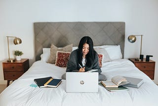 A person with a blanket draped around them sitting in bed working on a laptop, surrounded by books.