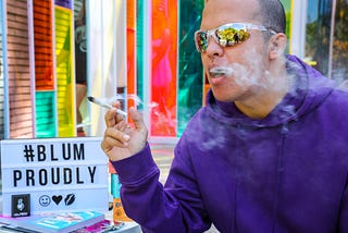 A person with a purple hoodie and sports sunglasses smoking a joint outside the Blüm storefront. #BlumProudly