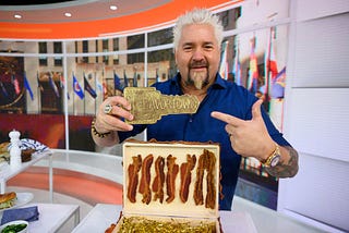 Guy Fieri holding and pointing to a small plaque that says “Flavortown”.