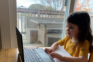 A young girl in a yellow dress sits at a table with a computer.