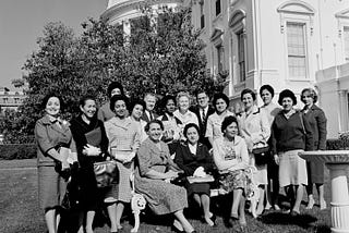 black-and-white photo of 17 people, the Latin American women’s group, posing outdoors in the East Wing Garden, 1961, showing the White House in the background