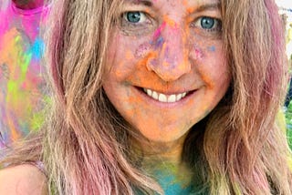 Midshot selfie of the female author smiling as she’s completely covered in the Holi Festival of Colors rainbow powders.