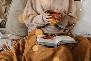 A woman is wearing a jumper and holding a hot drink. She has a blanket and a book on her lap.