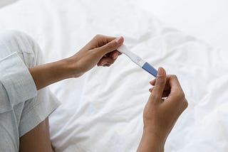 Close-up of a person holding a positive pregnancy test, showing two lines, while sitting on a bed.