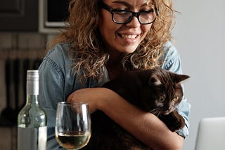 A woman with curly hair and glasses leans over a computer, smiling while holding a cat, with a bottle and glass of wine