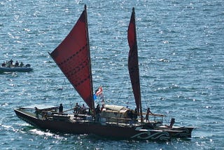 The waka Haunui floating on a rippled ocean with a small inflatable boat in the background. The waka is a replication of the style of canoe that early Māori may have used to voyage to Aotearoa New Zealand.