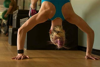 A woman is on her hands in a half handstand, with her feet up on a block behind her. She is in the middle of a pushup.