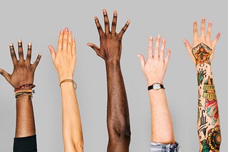 Six arms and hands raised up as if to volunteer, multiple ethnicities, set against a grey background.