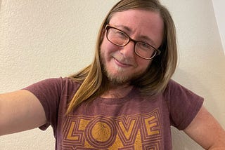 The author has long brown hair, glasses, a beard, a mustache, and is wearing a t-shirt that says “Love Y’all”. He has a mischievous smile that shows more in his tired eyes.