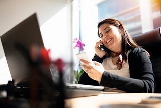 A photo of a smiling woman on the phone at her desk in front of her computer.