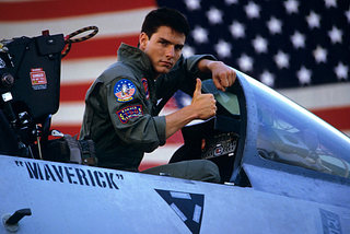 Maverick is standing in the cockpit of his jet looking straight into the camera. He has his right hand in a thumbs up sign. He is wearing Naval Aviator attire, which is a dark grey; there are Naval patches on it. Written on the side of the jet is his call sign, Maverick. Behind him there is a blurred out American flag.