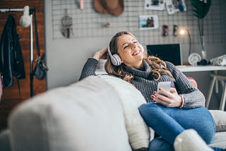 A woman holds her phone and smiles as she listens to a podcast on her headphones.
