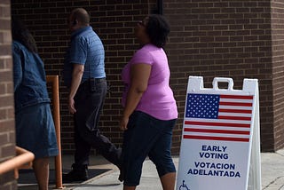 A photo of three early voters in Florida lining up at a poll. There is a “EARLY VOTING VOTACION ADELANTADA” sign.
