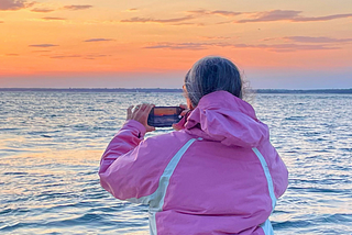 Middle aged woman wearing a pink wind breaker, on a boat, seen from behind. She is taking a picture of sunset with her phone.