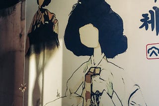 A wall at an angle covered with a large decal of a traditionally dresses Korean woman with large triangle shaped hair and no face. A smaller, similar woman is to the upper left of the photo and Hangeul characters are halfshown on the right side.