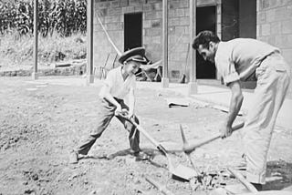 Black and white photo showing a bungalow under construction in the background; in the foreground, a child and adult playfully working the grounds with a shovel and pitchfork. Child is clearly having fun.