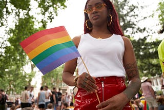 A young black woman holds up a rainbow flag at an outdoor gathering.