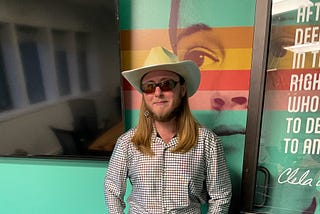 The author is wearing an opal cowboy hat with sunglasses, an opal shirt, blue jeans, and six-inch opal platform boots. The wall behind him is teal colored.