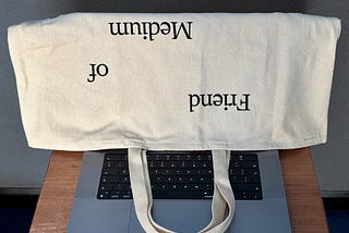 A laptop with the screen covered by an upside-down Friend of Medium tote bag.