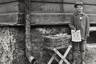 A sad-looking boy stands by a trestle table of newspapers, holding a copy for sale