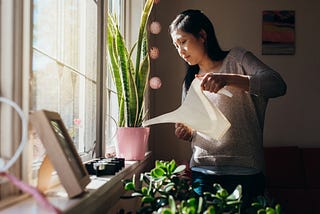 A photo of an Asian woman watering plants on her windowsill at home.