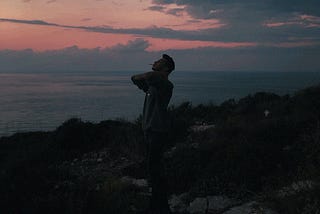 Male standing on a rocky beach just after sunset smoking a cigarette with hands clasped behind his neck