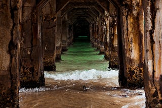 A photo taken underneath a dock, showing the waves of the sea crashing into the poles as it leads towards a dark end.