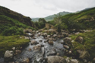 A stream running over rocks in a British moorland landscape. Green hills rising into cloudy skies.