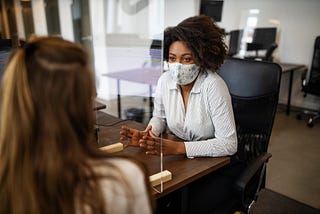 A Black businesswoman wearing a face mask negotiates with a client. There is a plastic divider for social distancing.