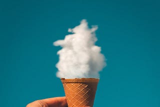 Ice cream cone being held up against a blue sky with a fluffy white cloud placed so it resembles ice cream in the cone.