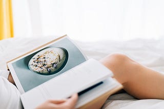 Closeup of a person, from the neck down, who’s reclining in bed, reading a cookbook with a large photo on one page.
