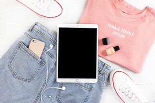 A folded “Female equals future” shirt, jeans with a phone in the back pocket, white canvas shoes, nail polish, and an iPad.