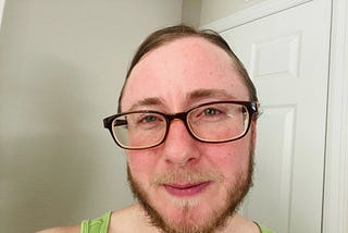 Photo of author with his hair pulled back, wearing glasses and a bright green sleeveless shirt. He looks peaceful. He has a beard and a mustache.