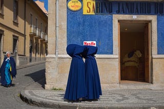 Two figures wearing blue robes stand together outside a building.