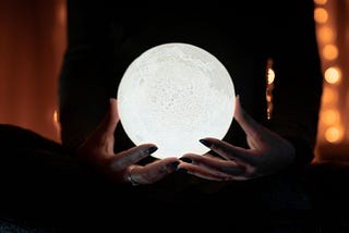 A silhouetted woman sits cross-legged, holding a bright white crystal ball.