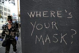 On the right side, three-quarters of the pic is a black exterior wall with “Where’s your mask?” written on it in chalk. The quarter of the photo on the left side is a person wearing latex gloves and a face mask, the latter under their chin.