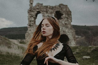 A woman with red hair wearing a black dress poses artfully with her eyes closed amid a backdrop of an overcast sky. There are brick ruins of a castle in the background and rolling hills.