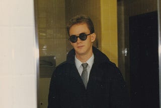 A sneering young man in a suit, black trenchcoat, and dark round sunglasses sneers into a mirror in a yellow-tiled restroom.