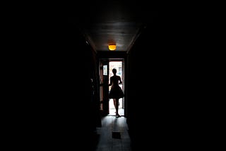 A mostly dark photo of a woman who is opening a door that casts light in the middle of the picture.