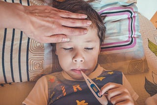 Child in bed with thermometer. Mom’s hand on his forehead
