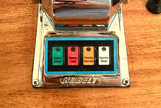 Colored closeup photo of a stainless steel sailboat drive shifter with four colored buttons marked left to right: (green) Station Select, (red) Station Lock, (orange0 Throttle Override, and (white) Synchro Mode