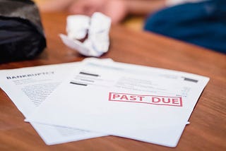 “Past Due” and bankruptcy notices on a table, with a despairing young man laying in the background.