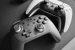A Nintendo Switch 2 Pro controller (left/bottom) and a white Sony DualSense controller sitting on a black desk mat.