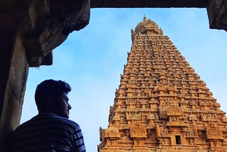 A man stands looking a temple structure in sand brown colour with blue sky behind.