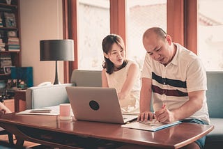 A couple sits on a couch in front of a laptop while one person writes in their notebook.