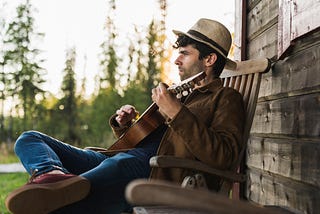Young man sitting on veranda of a wood house, playing the ukulele.