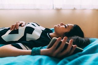 A young woman lies down and stares at the ceiling, holding her phone and feeling worried